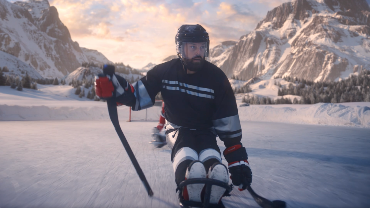 A Paralympic hockey player racing fiercely along ice with hockey stick in hand with a red hockey goal and snow-capped mountains in the background.